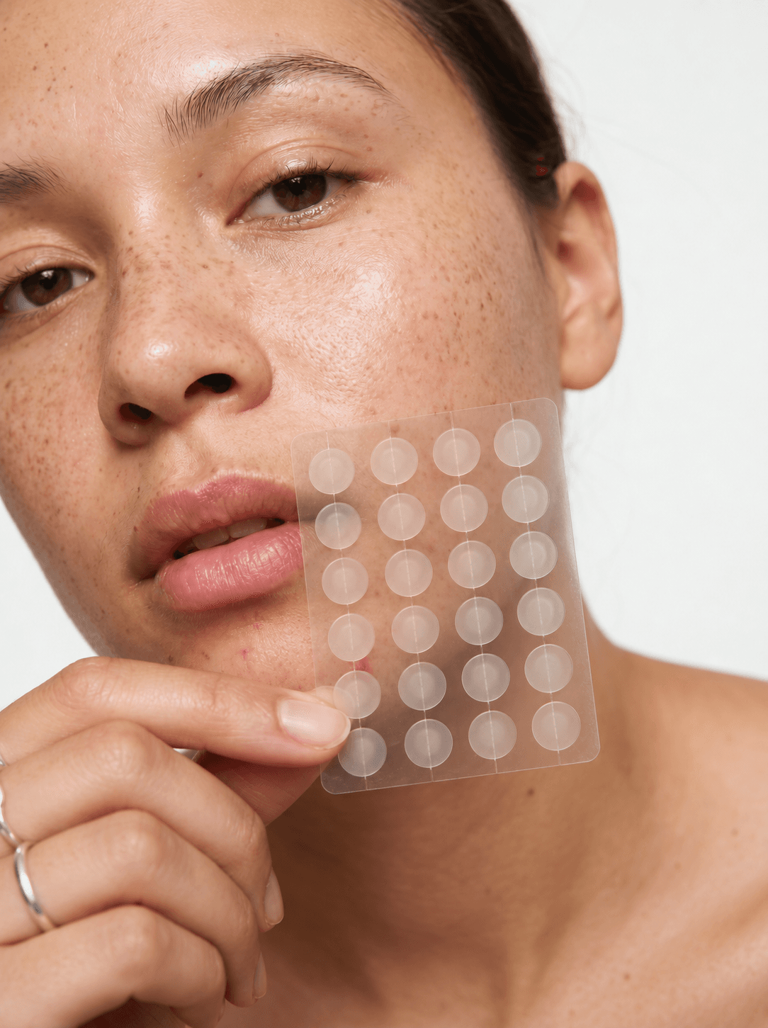 Woman holding a clear sheet of 24 MEND hydrocolloid pimple patches arranged in a four-by-six grid against her cheek